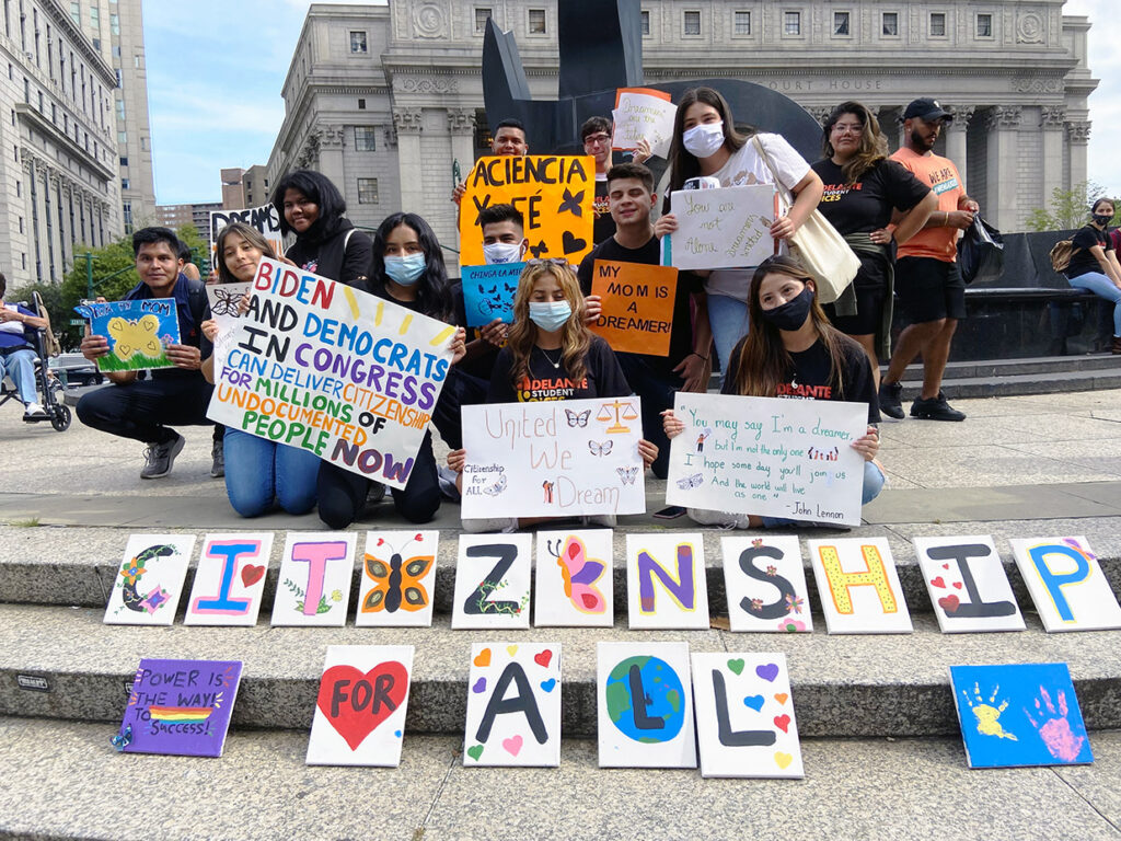 Latina/Latino youth stand and kneel together with handmade signs supporting immigration form. In front of them is a colorful sign saying, "Citizenship for All"