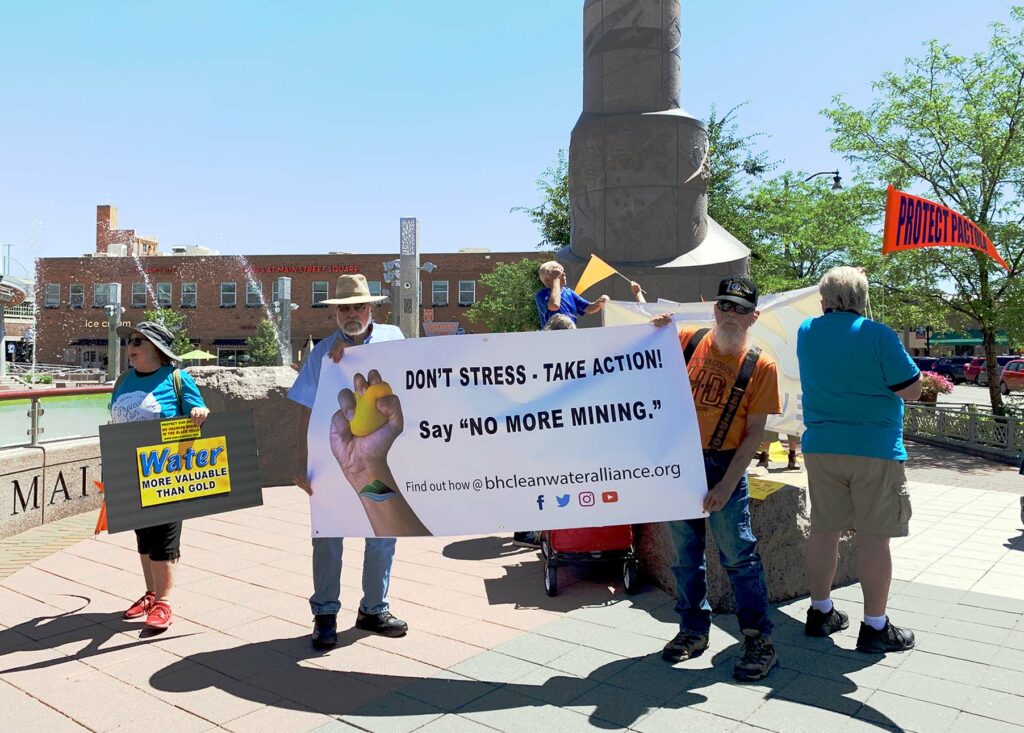 A group of older folks appear to be protesting near a community square by a fountain. Two older men hold a banner that reads: Don't Stress - Take Action! Say "NO MORE MINING"