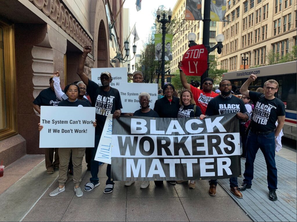 A group of black workers and white allies stand on a city sidewalk with signs and a banner that reads: Black Workers Matter