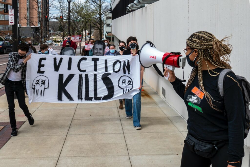 A group of people walk along a sidewalk holding a white banner with black painted words across that reads: Eviction Kills. There are cartoon images of skulls on the banner. A protester leads the group holding a loudspeaker.