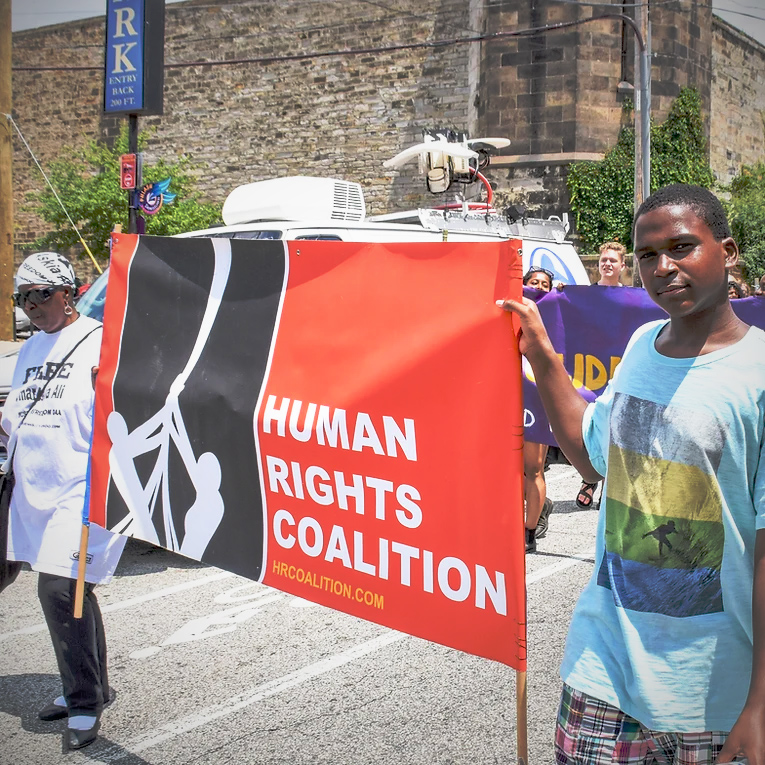 Within what looks like a march or protest, a young black male holds a red and black banner with an older black woman. The banner reads: Human Rights Coalition.